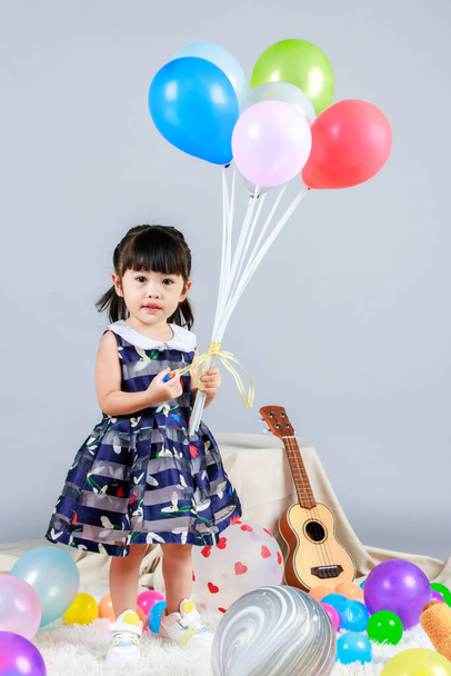 Küçük, masum anaokulu anaokulu kızı, mavi bir elbise giymiş, gri arka planda renkli balonlar tutan kameraya gülümseyen bir kızın portresi.. - Fotoğraf, Görsel