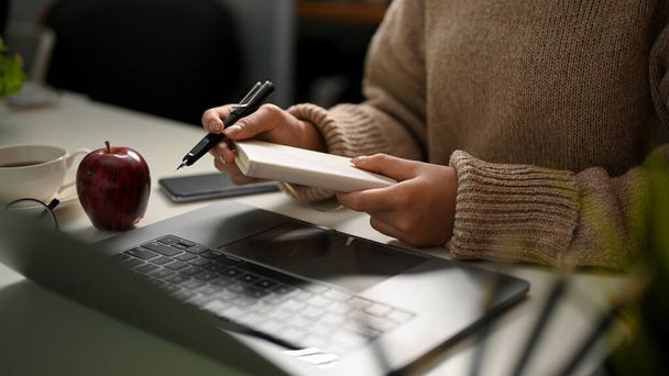 Przycięty obraz, kobieta w przytulnym swetrze robiąca notatki na swoim spiralnym notatniku, robiąca ważne listy na papierze przy biurku. - Zdjęcie, obraz