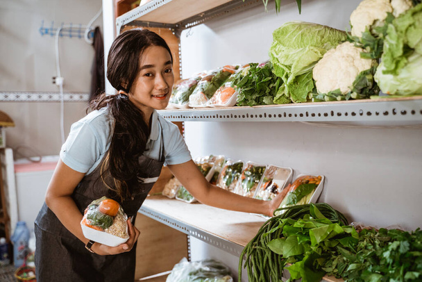 Smiling girl wearing apron arranging fresh vegetables on display case - 写真・画像