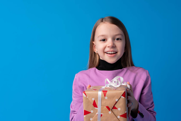 Happy little girl with long hair holding a gift box on a blue background - Fotoğraf, Görsel