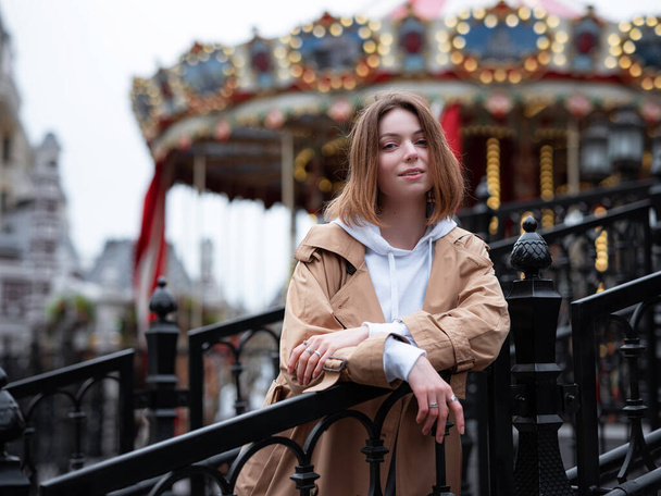 Beautiful woman in beautiful beige coat walk in autumn cloudy Moscow, enjoys and has fun on carousel. Street style. Travel to Russia concept. - Photo, Image