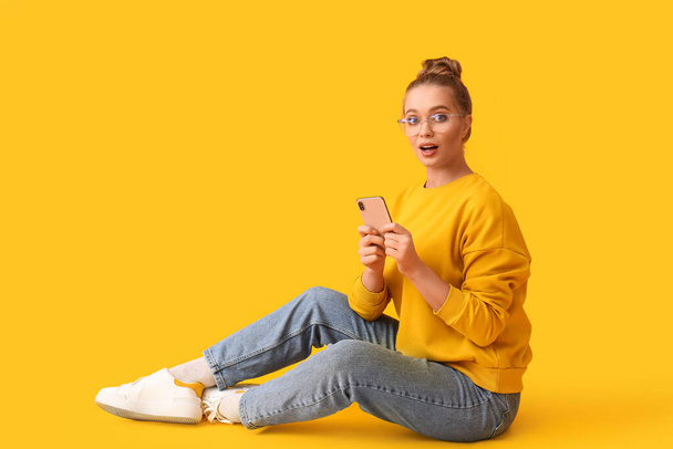 Здивована молода жінка з мобільним телефоном на жовтому фоні
 - Фото, зображення