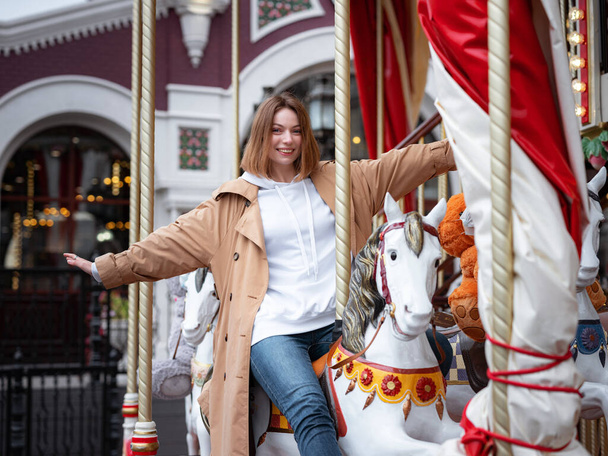 Beautiful woman in beautiful beige coat walk in autumn cloudy Moscow, enjoys and has fun on carousel. Street style. Travel to Russia concept. - Фото, зображення