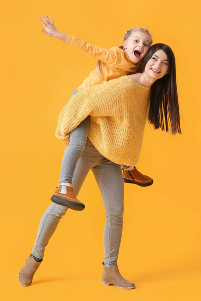Young mother carrying her little daughter piggyback on yellow background - Photo, Image