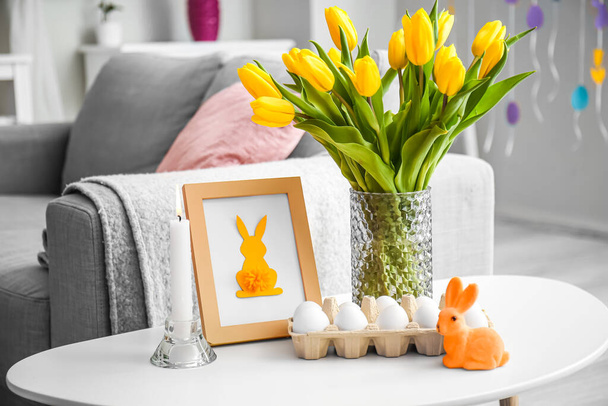 Suporte com ovos de Páscoa, moldura da foto, vaso com tulipas e vela acesa na sala de estar - Foto, Imagem