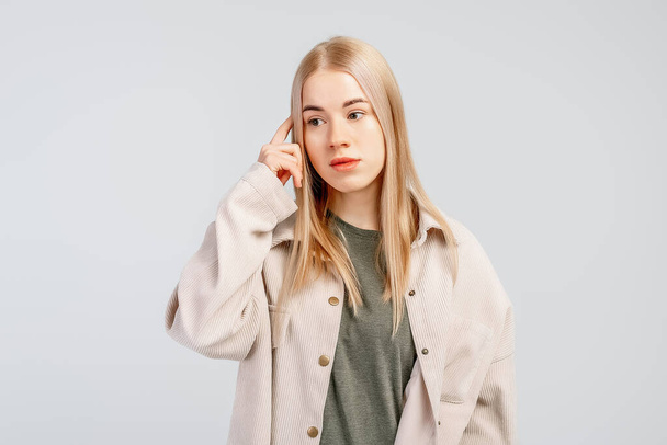 Image of confused fair-haired young woman thinking with finger to her temple, looking complicated, wears casual clothes, stands against grey background. Body language concept - Фото, изображение