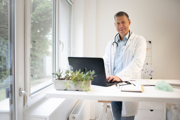 handsome doctor in white coat and blue shirt and stethoscope stands behind high table and works on laptop and has web conference or meeting - Фото, изображение