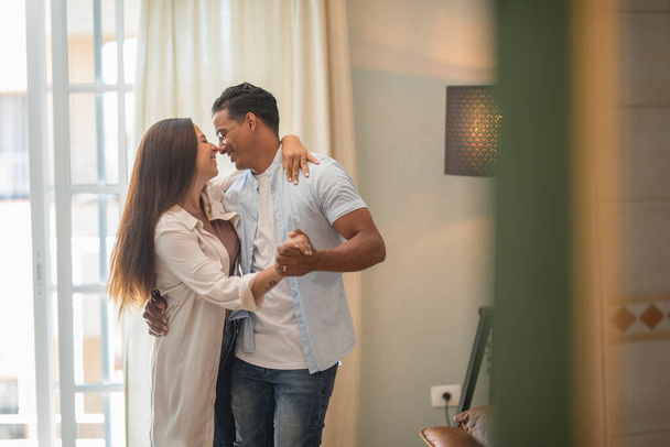 Interracial couple dance at home together having fun and love. New house owner and loan leisure indoor activity. Boy and girl in relationship have fun at home dancing and smiling to each other - Foto, immagini