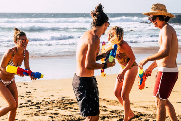 Szczęśliwi turyści bawią się na plaży bawiąc się pistoletami wodnymi. Młodzi mężczyźni i kobiety cieszą się wakacyjnymi wakacjami śmiejąc się razem w przyjaźni. Ocean i brzeg w tle - Zdjęcie, obraz