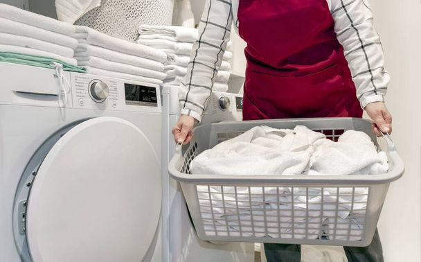 Pracownik w hotelu trzyma wiele ręczników w rękach w pobliżu pralki w pralni. - Zdjęcie, obraz