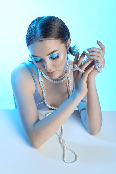 Beauty portrait woman makeup blue arrows eyes, beads jewelry around her neck, blue dress, woman at table with milk. Art makeup Professional makeup and cosmetics face. Sophisticated art portrait style - Foto, imagen
