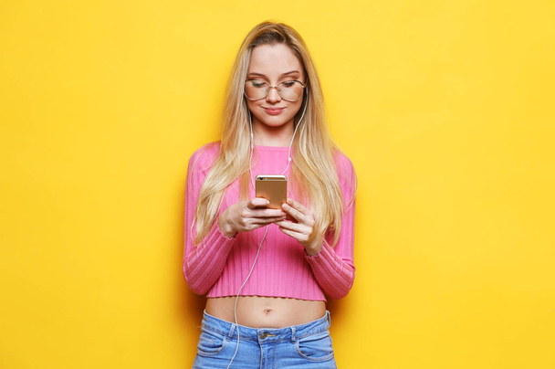Blond pretty woman in pink sweater smiling using smartphone posing on yellow background isolated smiling having fun, emotional - Foto, afbeelding