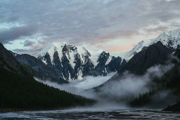 Scenic landschap met grote besneeuwde bergen op zonsondergang en dichte lage wolken in de bergvallei met bos silhouet. Sfeervol landschap met hoge besneeuwde berg bij zonsopgang en dikke lage wolken - Foto, afbeelding