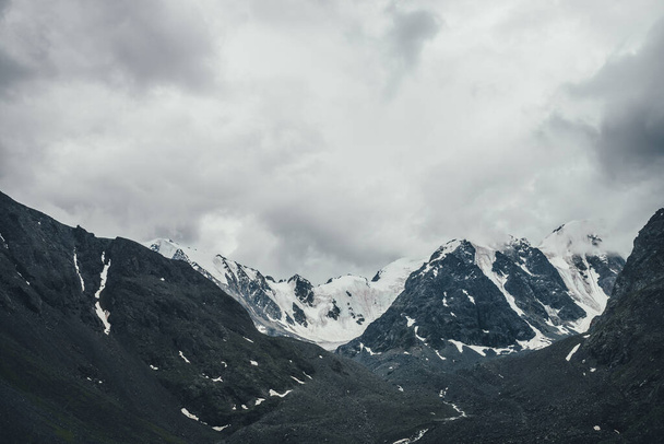 Σκούρο ατμοσφαιρικό ορεινό τοπίο με παγετώνα σε μαύρα βράχια σε γκρι συννεφιασμένο ουρανό. Χιονισμένα βουνά σε χαμηλά σύννεφα σε βροχερές καιρικές συνθήκες. Θολωμένο ορεινό τοπίο με μαύρα βραχώδη βουνά με χιόνι. - Φωτογραφία, εικόνα