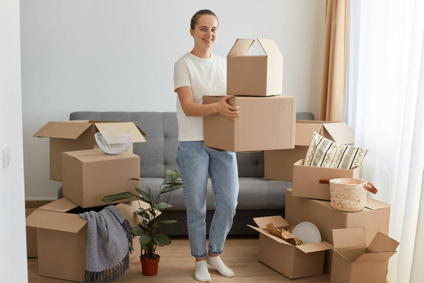 Full length portrait of Caucasian woman wearing white t shirt and jeans standing with cardboard box, looking at camera, unpacking personal pile during relocation into a new flat. - Foto, imagen