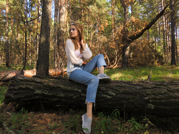 Young beautiful blonde woman in a silk blouse, white bra and blue jeans in the autumn forest on a fallen tree trunk - Фото, зображення