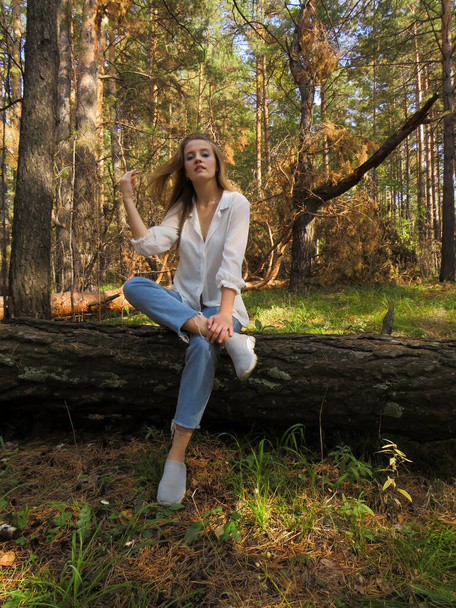 A young blonde woman in a white shirt and blue jeans in the autumn forest on a fallen tree trunk enjoys a vacation in nature, the nature of zabadnaya Siberia, the concept of outdoor activities, Hiking in the forest, travel and tourism - Foto, afbeelding