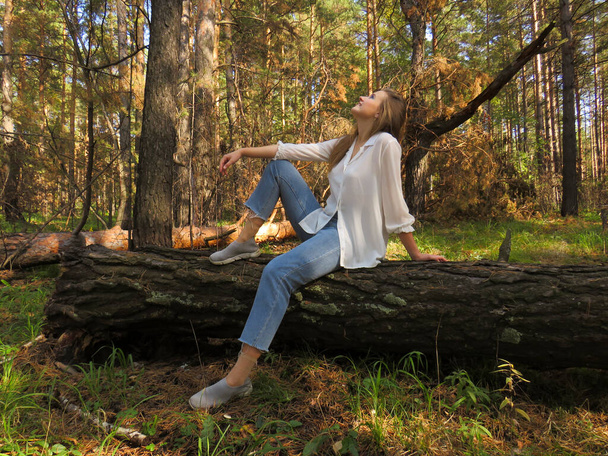 A young blonde woman in a white shirt and blue jeans in the autumn forest on a fallen tree trunk enjoys a vacation in nature, the nature of zabadnaya Siberia, the concept of outdoor activities, Hiking in the forest, travel and tourism - Foto, imagen
