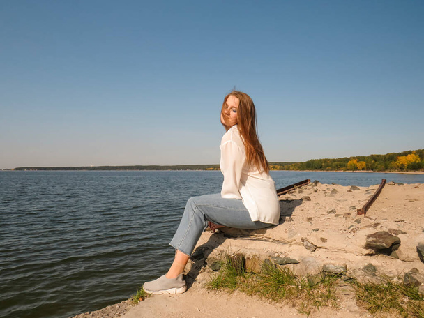 Blonde young woman in white silk blouse and blue denim pants on the old pier on the background of clear sky and blue waters of the sea bay enjoys colorful views of the landscape and outdoor recreation on a sunny autumn day - Foto, immagini