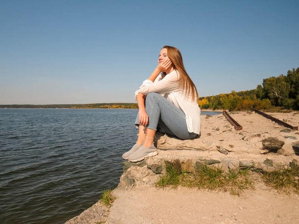 Blonde young woman in white silk blouse and blue denim pants on the old pier on the background of clear sky and blue waters of the sea bay enjoys colorful views of the landscape and outdoor recreation on a sunny autumn day - Foto, Bild
