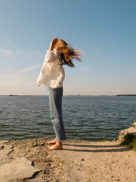 A barefoot blonde young woman in a white silk shirt and blue denim pants on an old stone pier against the background of the water of the sea bay enjoys a rest in nature on a clear sunny day - Photo, Image