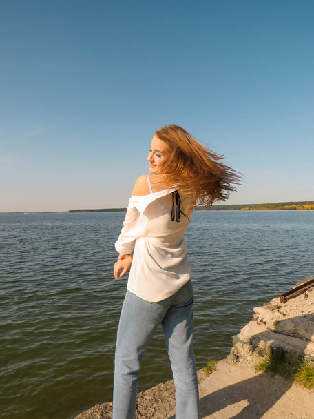 A barefoot blonde young woman in a white silk shirt and blue denim pants on an old stone pier against the background of the water of the sea bay enjoys a rest in nature on a clear sunny day - Φωτογραφία, εικόνα