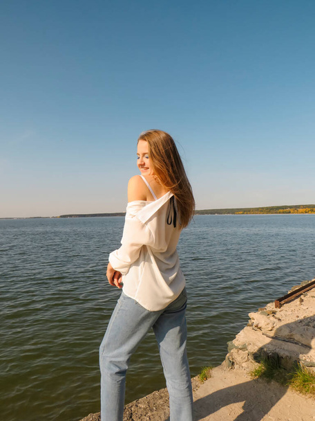 A barefoot blonde young woman in a white silk shirt and blue denim pants on an old stone pier against the background of the water of the sea bay enjoys a rest in nature on a clear sunny day - Foto, Imagem