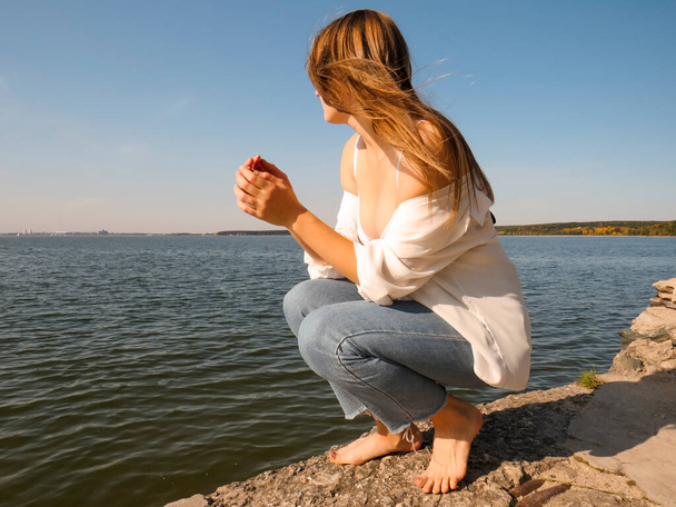 A barefoot young woman with blonde hair, in a white open shirt, a white bra on a beautiful chest and denim pants on an old pier on the seashore against the background of the water surface and blue sky on a sunny autumn day  - Foto, Imagem