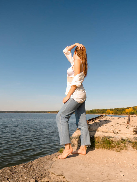 A barefoot young woman with blonde hair, in a white open shirt, a white bra on a beautiful chest and denim pants on an old pier on the seashore against the background of the water surface and blue sky on a sunny autumn day  - Фото, изображение