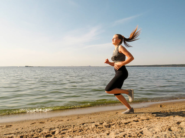 A young slender woman with beautiful long legs in black training tights and a white T-shirt running along the shore along the water's edge on a warm sunny autumn day. Concepts of outdoor sports, active lifestyle, sports and wellness running - Foto, Bild
