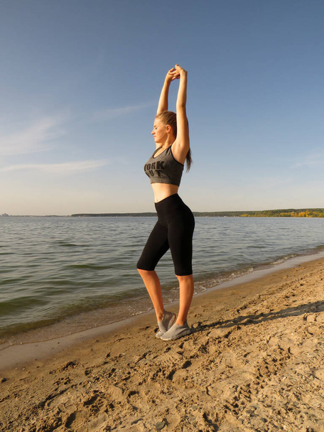 A slender young blonde woman with a sexy figure in sports shorts and a T-shirt does yoga in the fresh air on the seashore on a sunny warm day.Concept of outdoor sports, healthy lifestyle, outdoor activities - Photo, Image