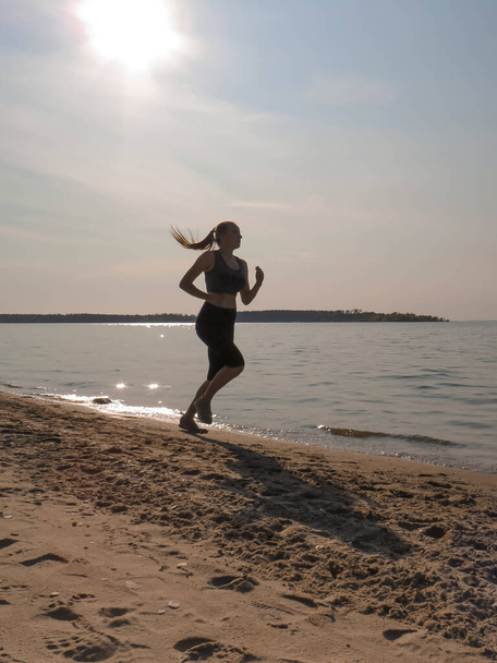 A young slender woman with beautiful long legs in black training tights and a white T-shirt running along the shore along the water's edge on a warm sunny autumn day. Concepts of outdoor sports, active lifestyle, sports and wellness running - Foto, Imagem