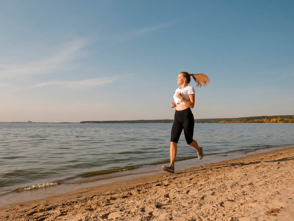 A young slender woman with beautiful long legs in black training tights and a white T-shirt running along the shore along the water's edge on a warm sunny autumn day. Concepts of outdoor sports, active lifestyle, sports and wellness running - Foto, Bild
