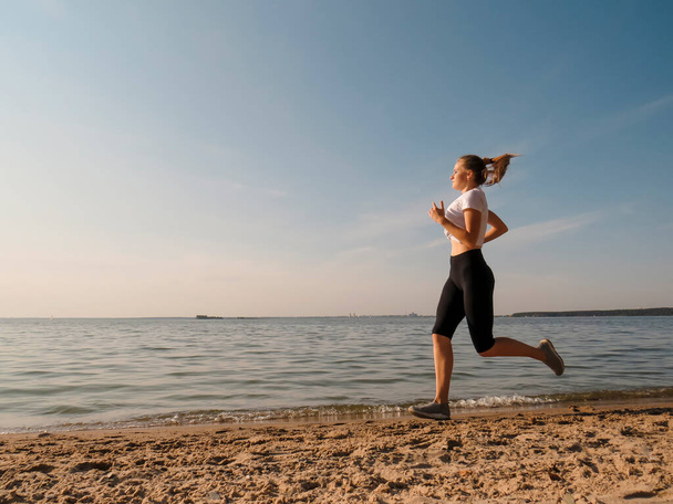 A young slender woman with beautiful long legs in black training tights and a white T-shirt running along the shore along the water's edge on a warm sunny autumn day. Concepts of outdoor sports, active lifestyle, sports and wellness running - Фото, зображення