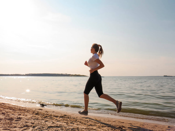 A young slender woman with beautiful long legs in black training tights and a white T-shirt running along the shore along the water's edge on a warm sunny autumn day. Concepts of outdoor sports, active lifestyle, sports and wellness running - Фото, изображение