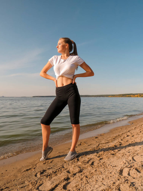 A slender young blonde woman with a sexy figure in sports shorts and a T-shirt does yoga in the fresh air on the seashore on a sunny warm day.Concept of outdoor sports, healthy lifestyle, outdoor activities, - Photo, Image