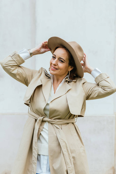 Woman in stylish trench coat and hat looking at camera while posing outdoors on the street. - Foto, afbeelding