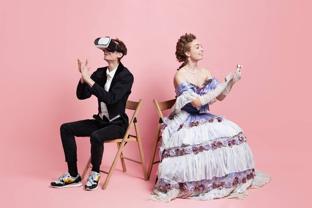 Two people young beautiful woman in image of medieval royal person in evening dress with servant, page-boy using gadgets isolated on pink background. - Photo, Image