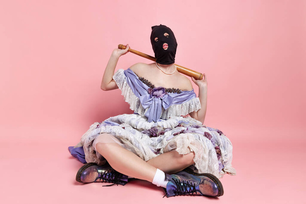 Comic portrait of young funny girl in image of medieval royal person in renaissance style dress and black balaclava isolated on pink background. Comparison of eras, beauty - Photo, image