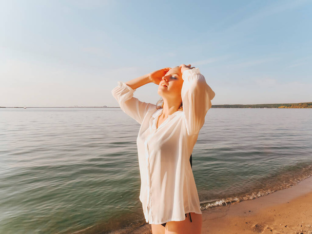 A happy young woman in a white shirt and bikini on a deserted seashore at sunset, peace and tranquility, the concept of outdoor recreation, beach sun tanning, active lifestyle, harmony and unity with nature, relaxing in silence in the fresh air - Photo, Image
