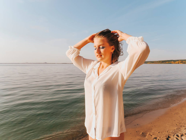 A happy young woman in a white shirt and bikini on a deserted seashore at sunset, peace and tranquility, the concept of outdoor recreation, beach sun tanning, active lifestyle, harmony and unity with nature, relaxing in silence in the fresh air - Фото, изображение