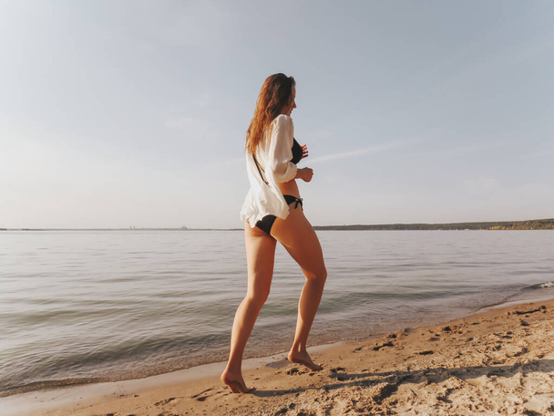 An attractive young woman in a black bikini and a silk shirt with tanned slender legs running along the seashore along the water's edge at sunset, the concept of outdoor recreation, sun tanning, active lifestyle, outdoor sports - Zdjęcie, obraz