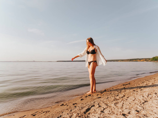 Attractive young blonde woman with a beautiful tanned body in a black bikini on a sandy beach at the water's edge takes a sun tan at the end of a hot day, the concept of the beach season, sun tanning and swimming, active lifestyle - Foto, Imagem