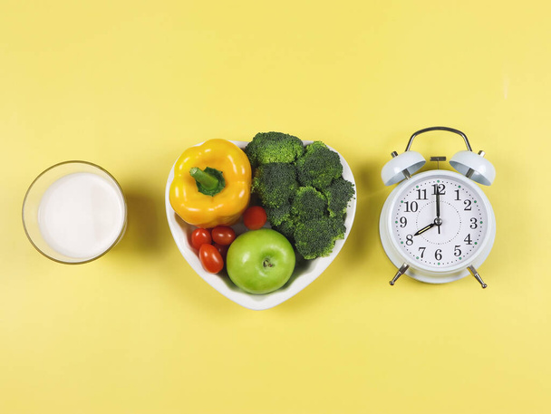 Top view of vegetables  capsicum, broccoli, tomatoes and apple  in heart shape plate, white  vintage alarm clock and a glass of milk  on yellow background with copy space .  healthy lifestyle  and intermitten fasting concept. - Фото, изображение