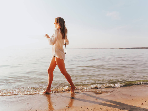 An attractive young woman in a black bikini and a silk shirt with tanned slender legs running along the seashore along the water's edge at sunset, the concept of outdoor recreation, sun tanning, active lifestyle, outdoor sports - Photo, Image