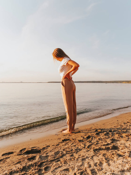 An attractive blonde young woman with bare feet on the sandy shore of the sea bay enjoys a sunset vacation, emotions of joy and freedom, concepts of unity and harmony with nature, outdoor recreation, lifestyle, active lifestyle, vacation at sea - Foto, imagen