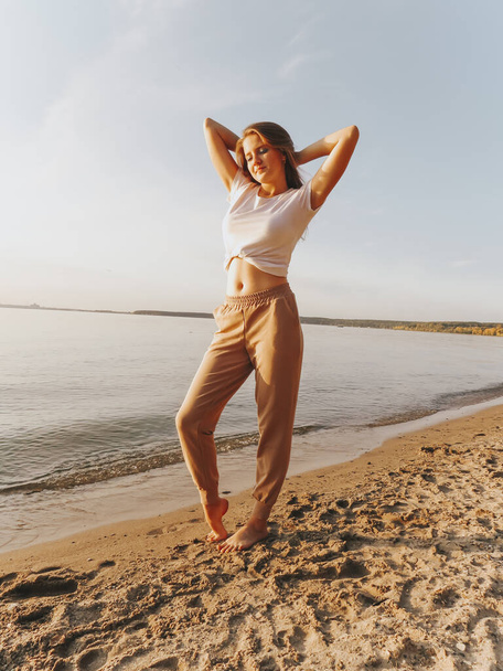 An attractive blonde young woman with bare feet on the sandy shore of the sea bay enjoys a sunset vacation, emotions of joy and freedom, concepts of unity and harmony with nature, outdoor recreation, lifestyle, active lifestyle, vacation at sea - Foto, Imagem
