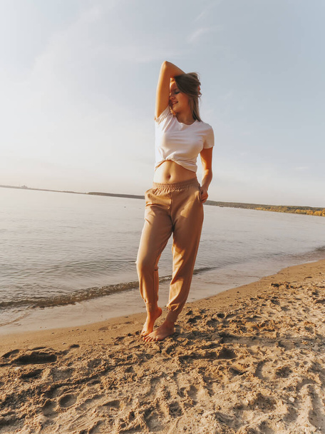 An attractive blonde young woman with bare feet on the sandy shore of the sea bay enjoys a sunset vacation, emotions of joy and freedom, concepts of unity and harmony with nature, outdoor recreation, lifestyle, active lifestyle, vacation at sea - Фото, зображення