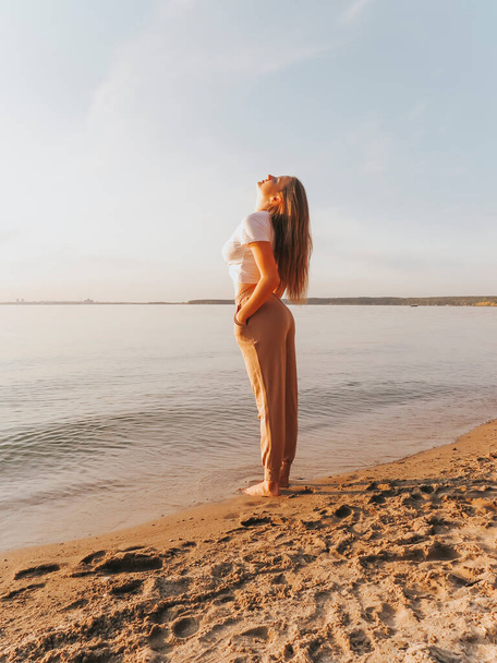 An attractive blonde young woman with bare feet on the sandy shore of the sea bay enjoys a sunset vacation, emotions of joy and freedom, concepts of unity and harmony with nature, outdoor recreation, lifestyle, active lifestyle, vacation at sea - Zdjęcie, obraz