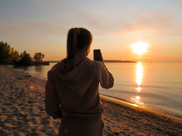 A young woman on a sandy sea beach at sunset takes a selfie on a mobile phone, the concept of travel and tourism, mobile technologies in travel, the beauty of natural landscapes, an active lifestyle, selfies for social networks - Fotó, kép